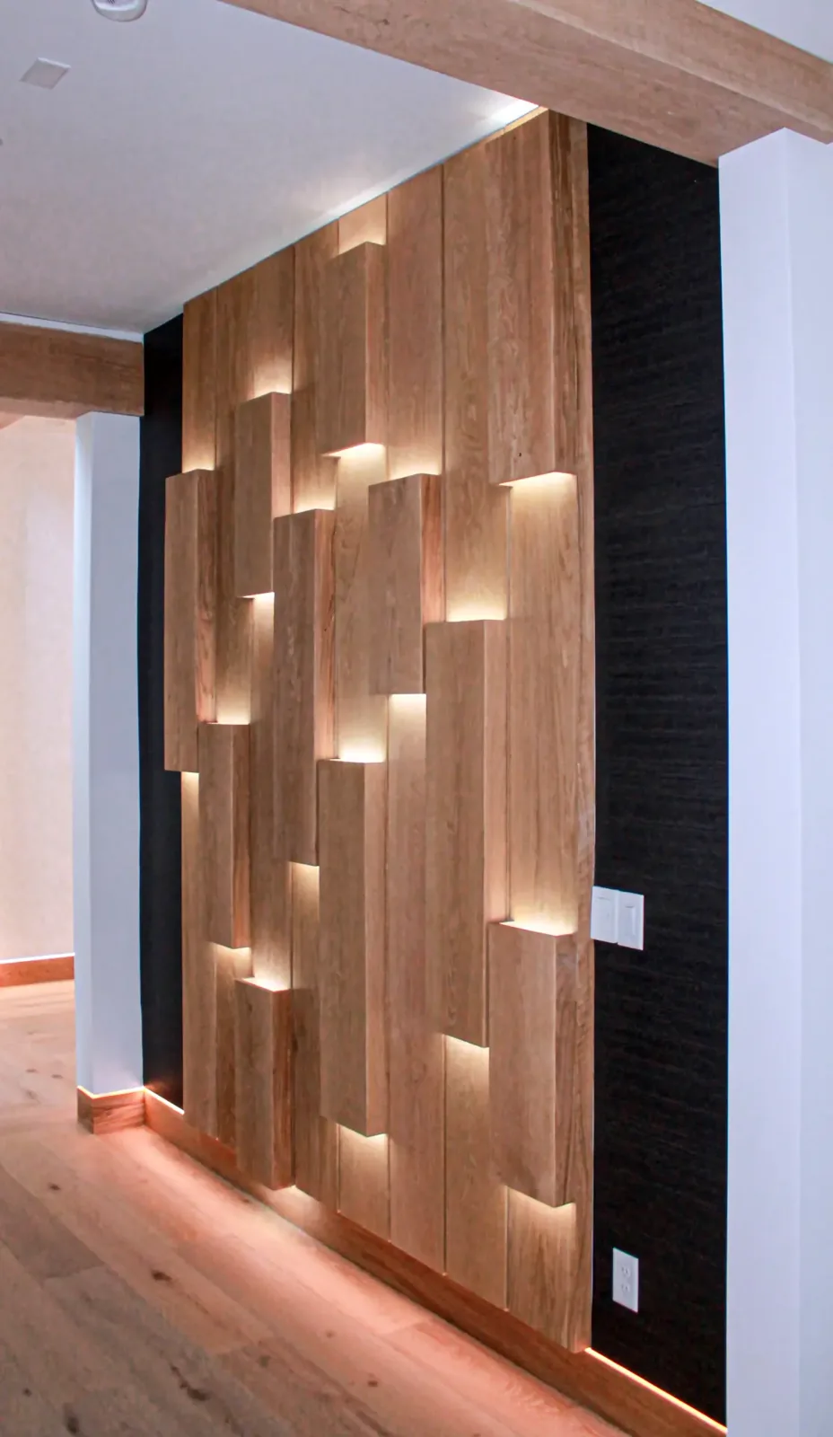 Backlit staggered wood plank wall seen from a hallway with a dark panel and light switches beside it